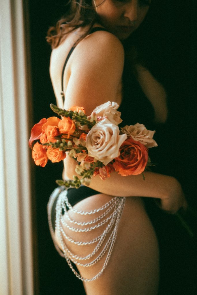 Delphine in a chain garment that covers her ass. She is holding a big boquet of orange and cream colored flowers. The garment highlights the curves of her 40 inch ass.