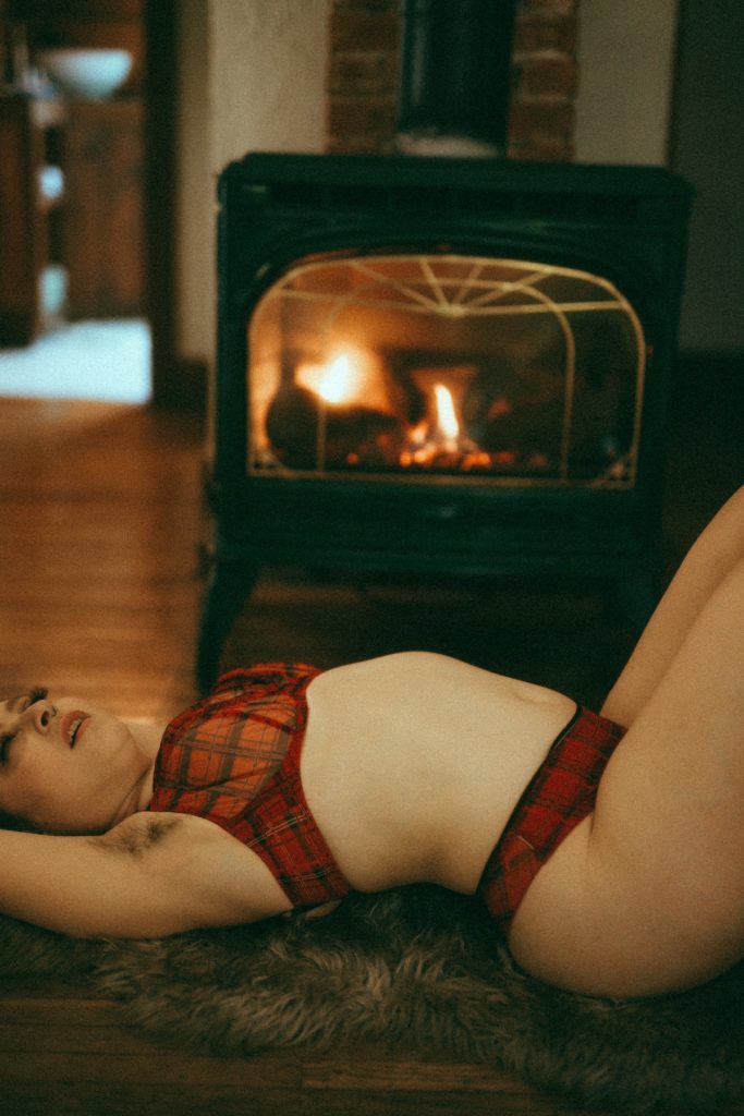 Delphine in plaid lingerie lying on her back in front of a fireplace. She is on a fur rug surrounded by books.