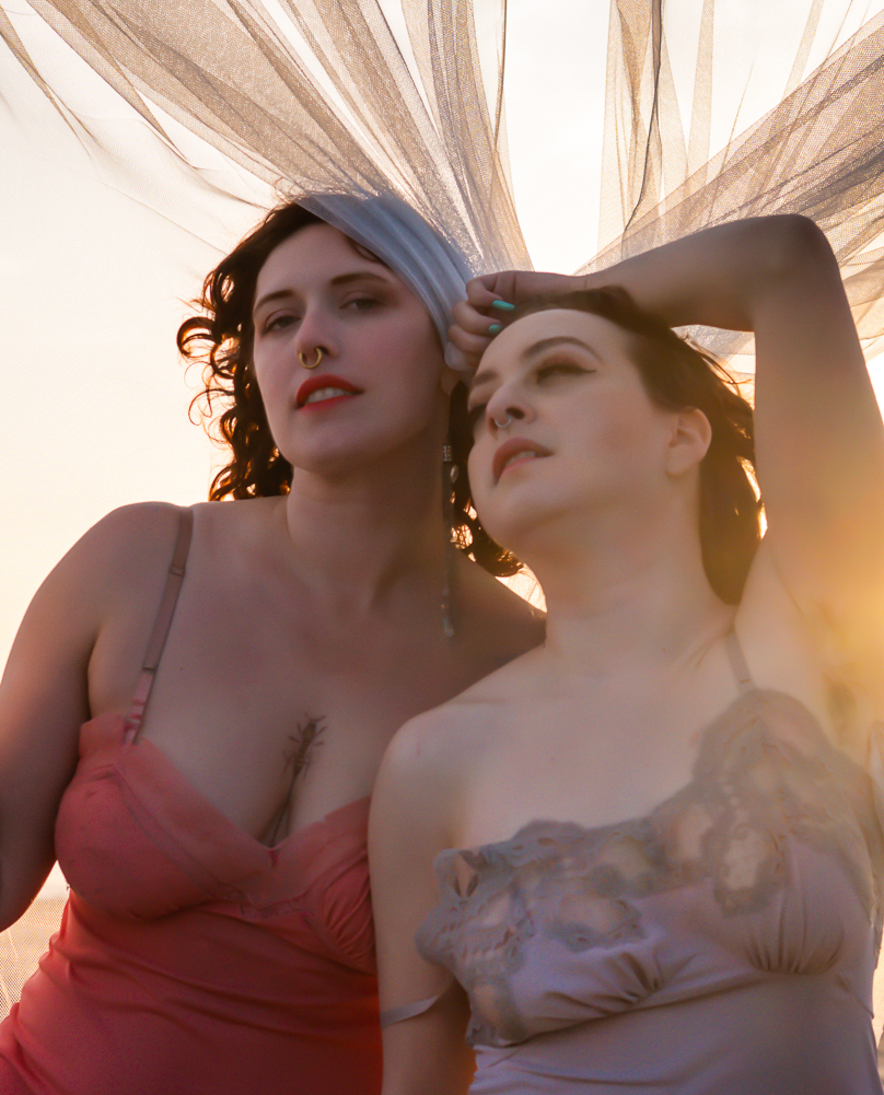 Delphine is a short brunette white woman with brown hair. She is on the right. She is wearing a mauve lace slip. She has her armpit raised to reveal her armpit hair. Flora is on the left. She is a taller white woman with red hair. She is wearing a coral slip. Delphine is staring wistfully in the distance and Flora is staring seductively at the camera. You can book a duo with these two SF Bay Area escorts.