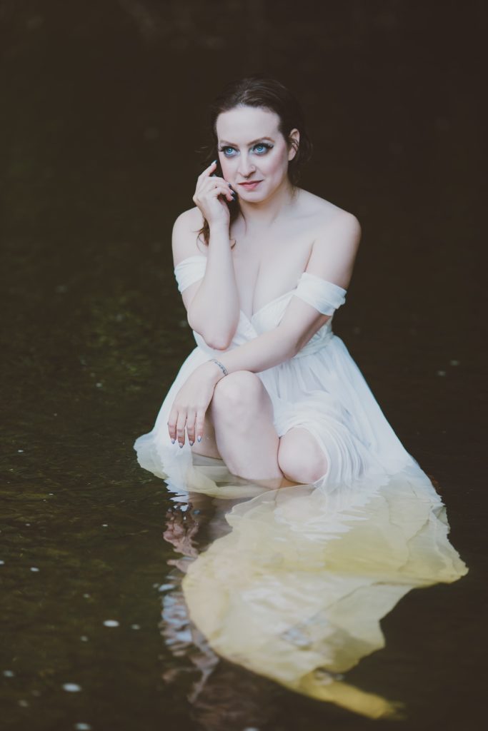 Delphine, a white woman with brown hair and big blue eyes, wearing a mythical white dress in a mysterious and murky body of water. 