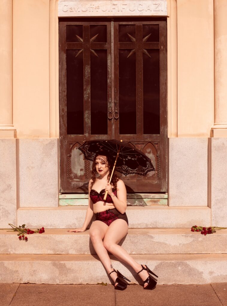 delphine, a brunette woman in maroon lingerie and black pleasers in front of a cemetery building.