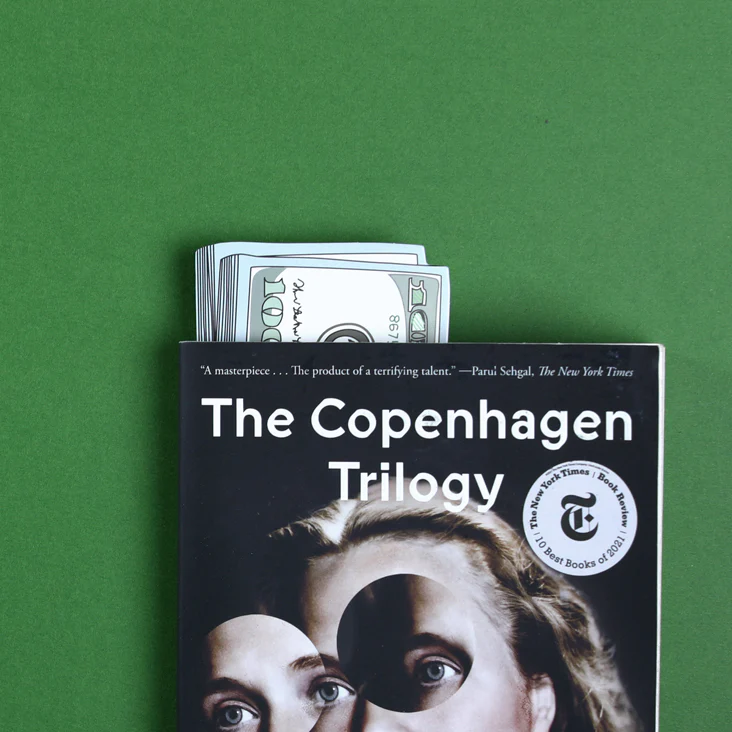 A book called The Copenhagen Trilogy with a bookmark that looks like a fake stack of $100 bills sticking out of it.