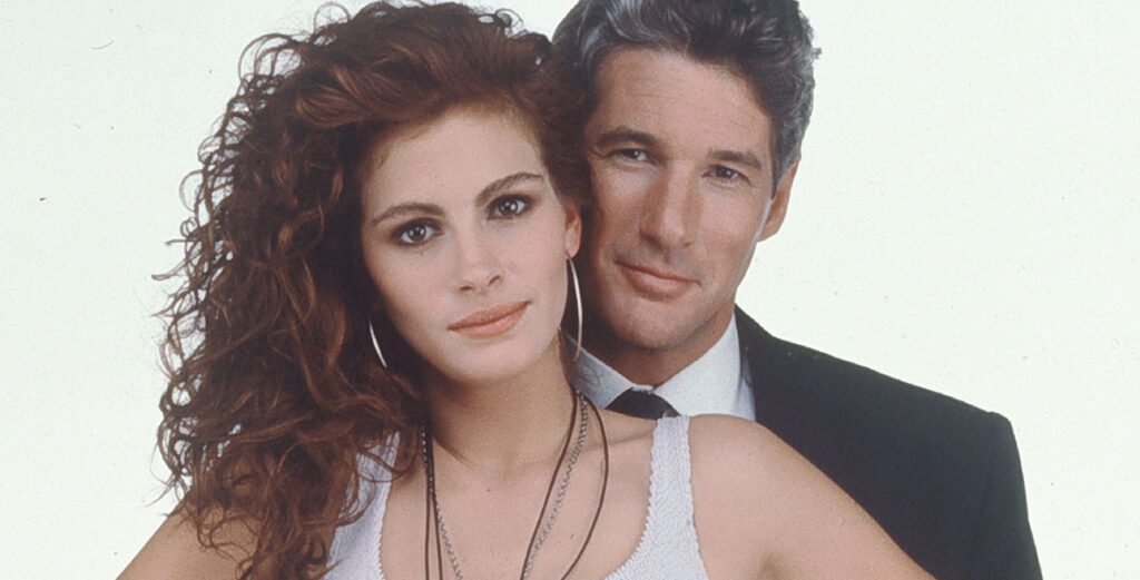 Julia Roberts a white woman with long curly brown hair wearing a white tank top and silver hoop earrings. She is standing in front of Richard Gere, and actor who is conventionally handsome with brown and silver hair. He is wearing a suit.