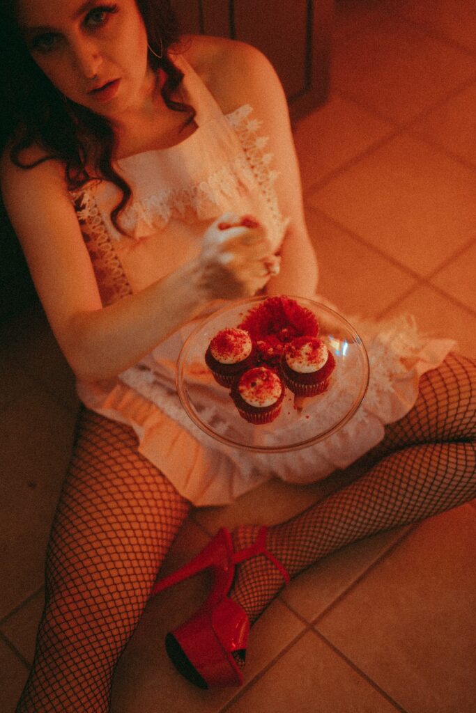 Delphine wearing a pink apron staring at the camera. She is smashing a red velvet cupcake. She is wearing a pink apron, red pleasers, and not much else. The vibe is a little Fiona Apple Criminal.