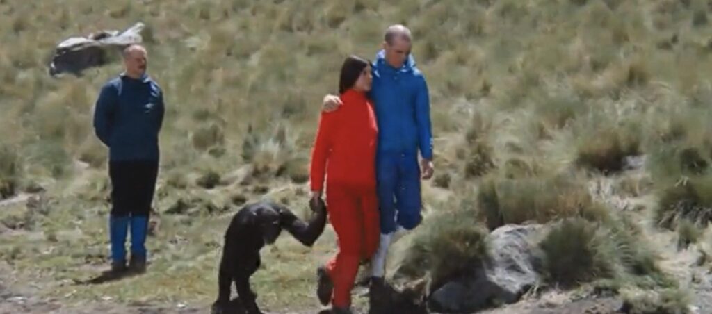 The Thief in a blue sweatsuit, and The Prostitute in a red one. She is holding the hand of a chimp. Alejandro Jodorowsky, The Alchemis is behind them. They are standing in a field.