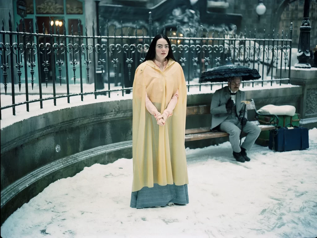 Emma stone as Bella Baxter in Poor Things. She is in a long yellow cape in a snowy scene. Behind her is Mark Ruffallo as her jilted lover. He is sitting on a park bench.