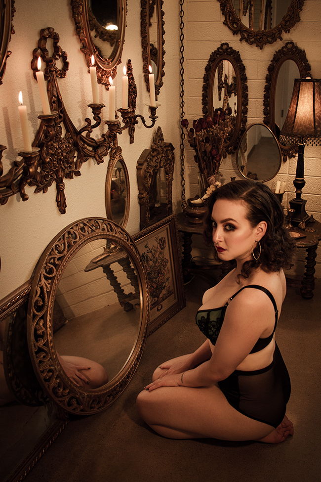 Delphine is in an ornate green bra and panty set. She is kneeling on the ground in front of some mirrors and a candlabra.