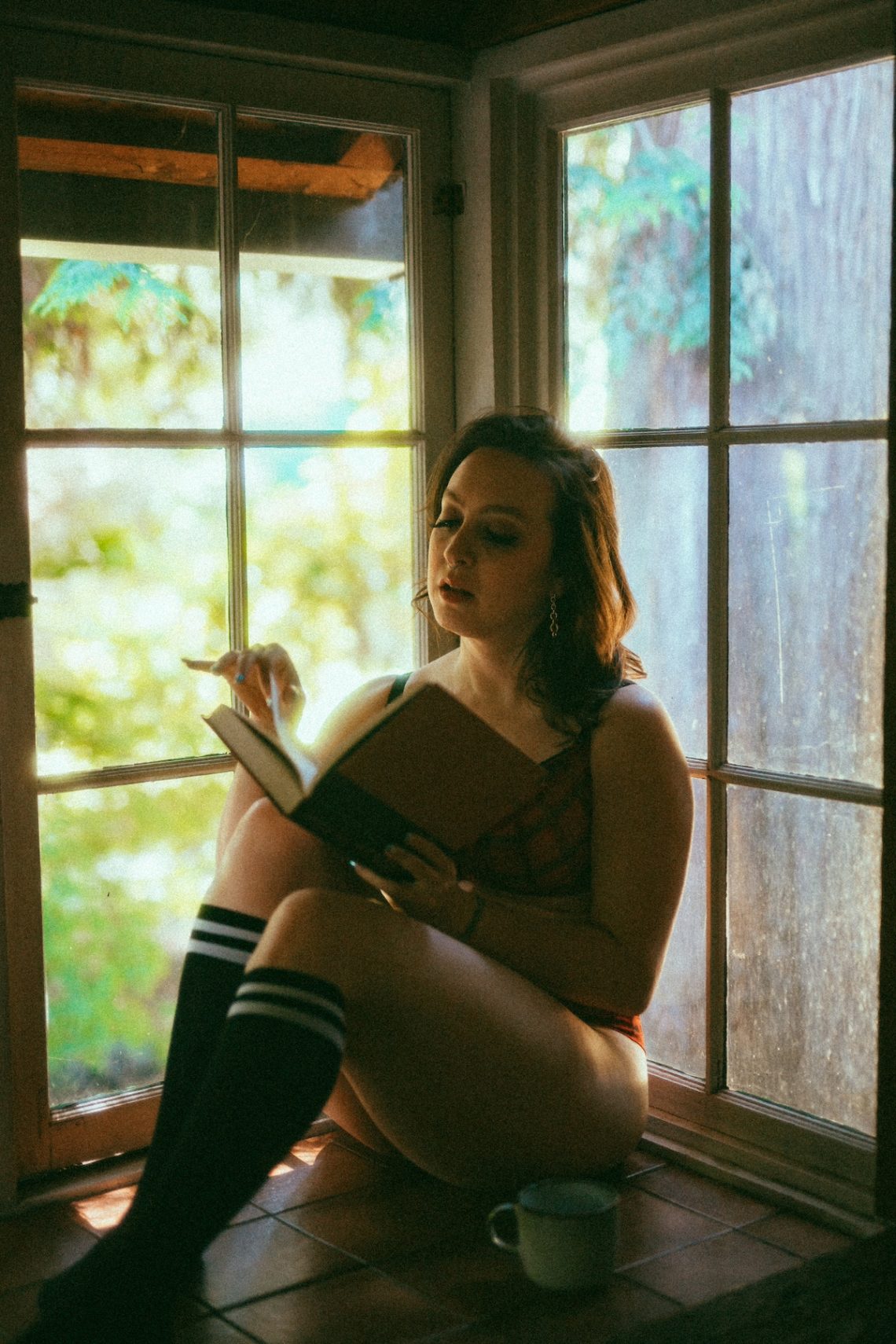 Delphine is sitting in a bay window reading a book. She is wearing lingerie and knee socks.