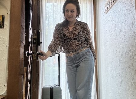 delphine brunette woman in leopard top and jeans opening the door next to a blue suitcase.