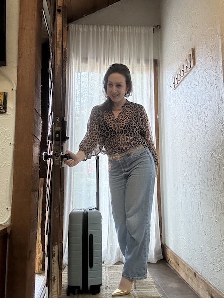 delphine brunette woman in leopard top and jeans opening the door next to a blue suitcase.