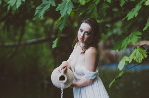 Delphine in a flowing white dress holding an ampoule. She is standing a river with green trees behind her. She is looking like Aquarius, the water bearer, or a siren who will lure you into the water.