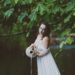 Delphine in a flowing white dress holding an ampoule. She is standing a river with green trees behind her. She is looking like Aquarius, the water bearer, or a siren who will lure you into the water.