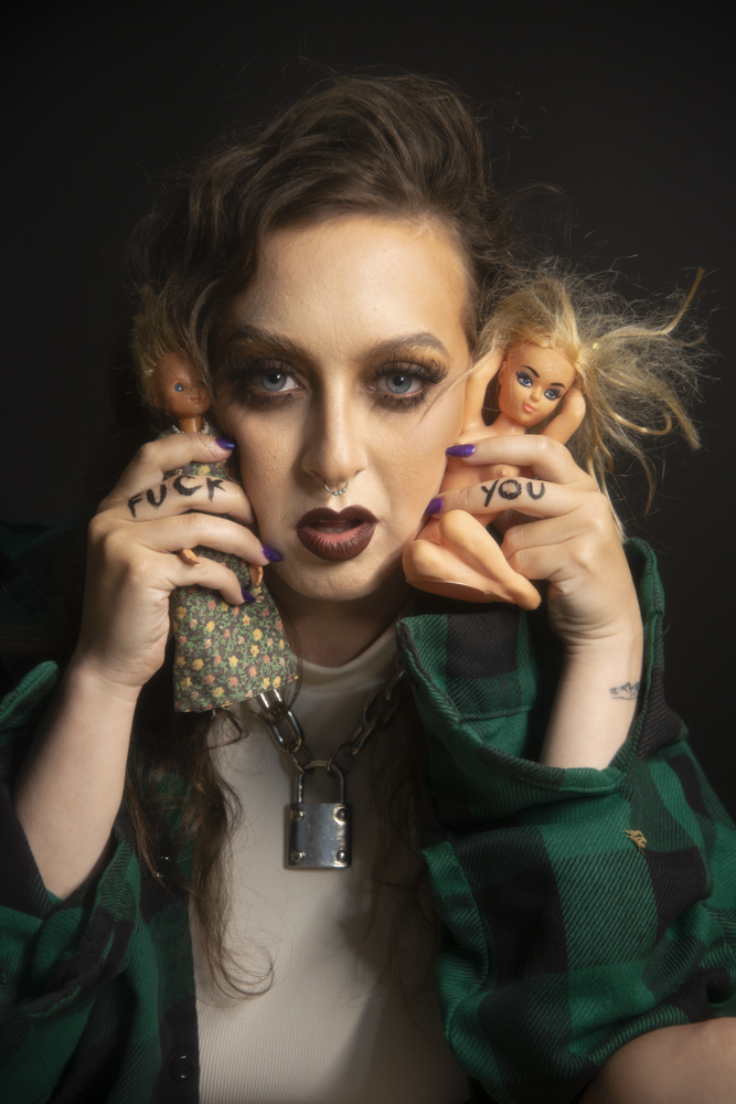 Delphine in a green plaid shirt and white tank top. She has a silver padlock necklace on. She is holding two creepy dolls in each hand. Her fingers have "fuck you" written on them. This photo is by Shannon Brooke.