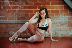 Delphine in a too-small sports bra that says Juicy on it, pink velour track pants, and white pleaser heels. She is wearing pink metal rim sunglasses and is posing on a red floor in front of a brick wall. This photo is by Shannon Brooke.