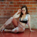 Delphine in a too-small sports bra that says Juicy on it, pink velour track pants, and white pleaser heels. She is wearing pink metal rim sunglasses and is posing on a red floor in front of a brick wall. This photo is by Shannon Brooke.