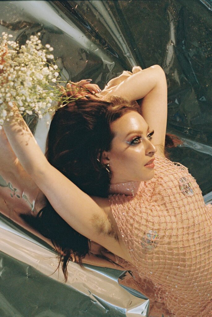 Delphine in a pink fishnet dress with silver pasties on underneath. Her arm is raised so you can see her armpit hair. She has a bundle of flowers in her hands. She is laying on a silver background. She is looking away from the camera as if she is staring at her beloved. This photo is by Shannon Brooke.