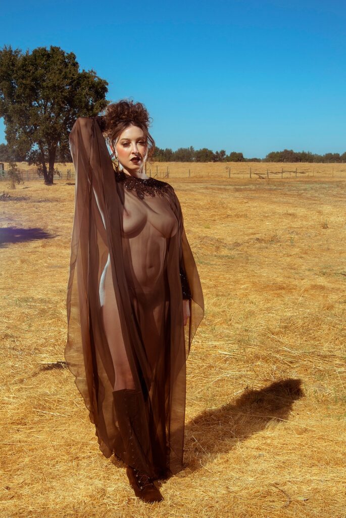 Delphine in a sheer brown mumu. She is standing in a dry field with an oak tree behind her. She looks like a farm witch.