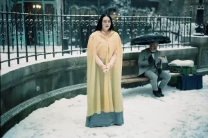 Emma stone as Bella Baxter in Poor Things. She is in a long yellow cape in a snowy scene. Behind her is Mark Ruffallo as her jilted lover. He is sitting on a park bench.