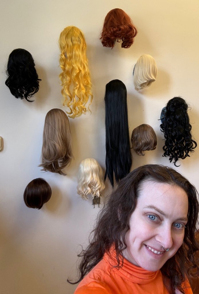 Delphine is wearing an orange turtlneck and no makeup. She is standing in front of a decorative wall with 10+ wigs of all lengths and colors.