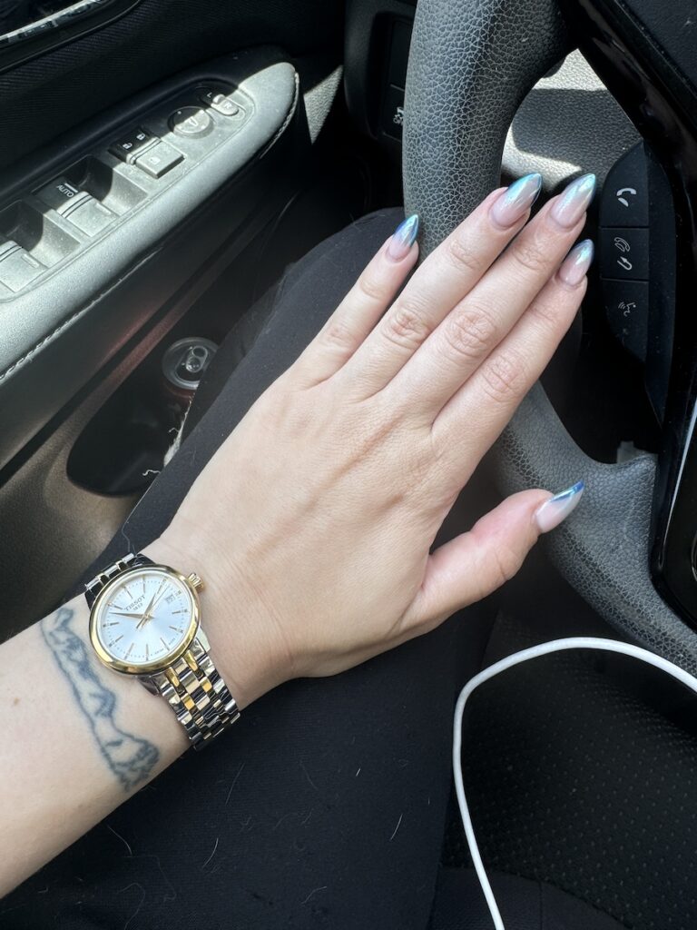 Close up of Delphine's nails in the car. They are cream with an icy blue shiny edge. She has on a silver and gold Tissot watch.