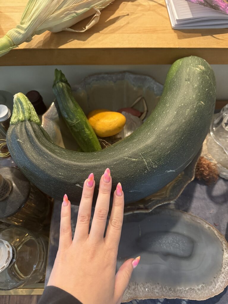 Delphine's hand with a giant zucchini. Her nails are neon tie dye colors.