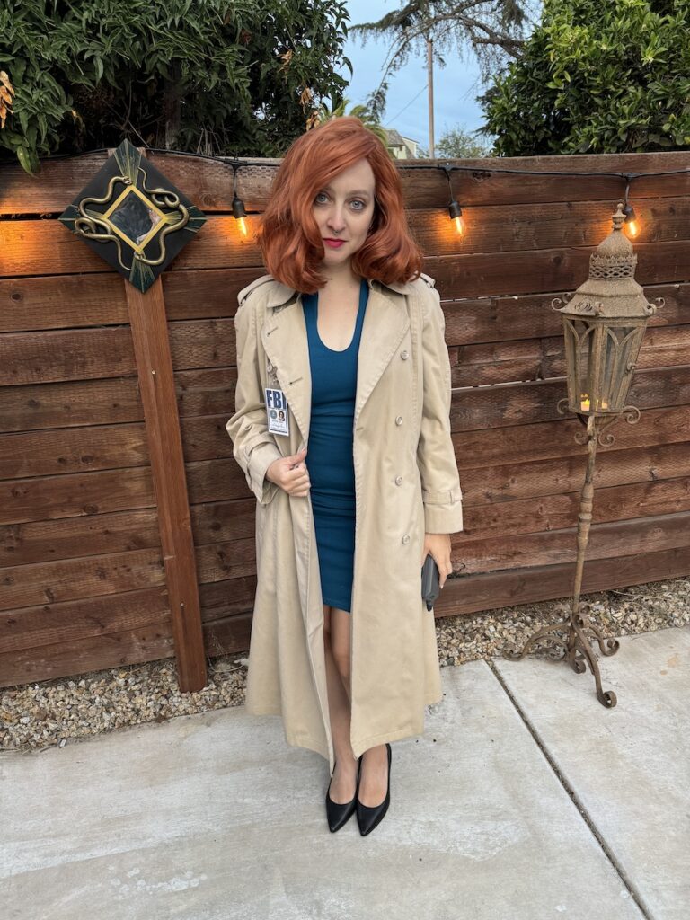 Delphine in a red wig and trench coat with a blue dress under it. her costume is Dana Scully from the X Files. She is standing in front of a wooden fence.