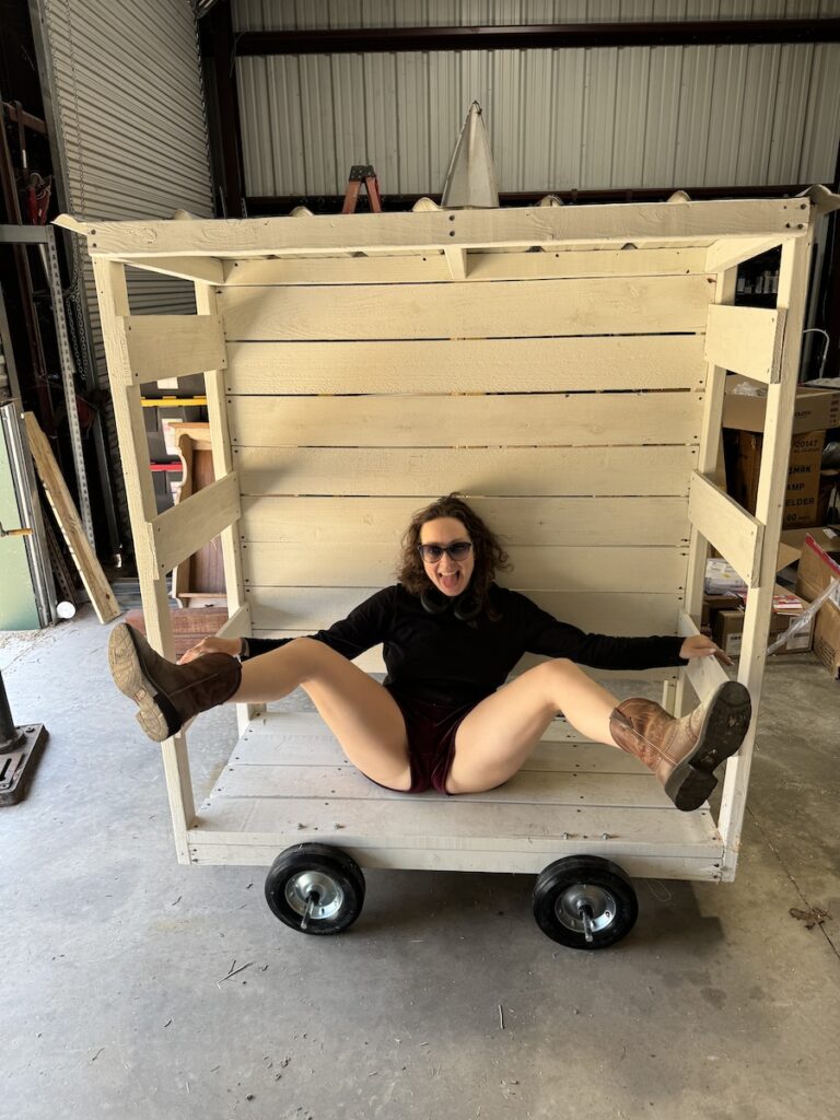 Delphine wearing black shorts, cowboy boots, and a long black shirt. She is sitting in a white cart that she built. Her legs are up in the air and spread wide.