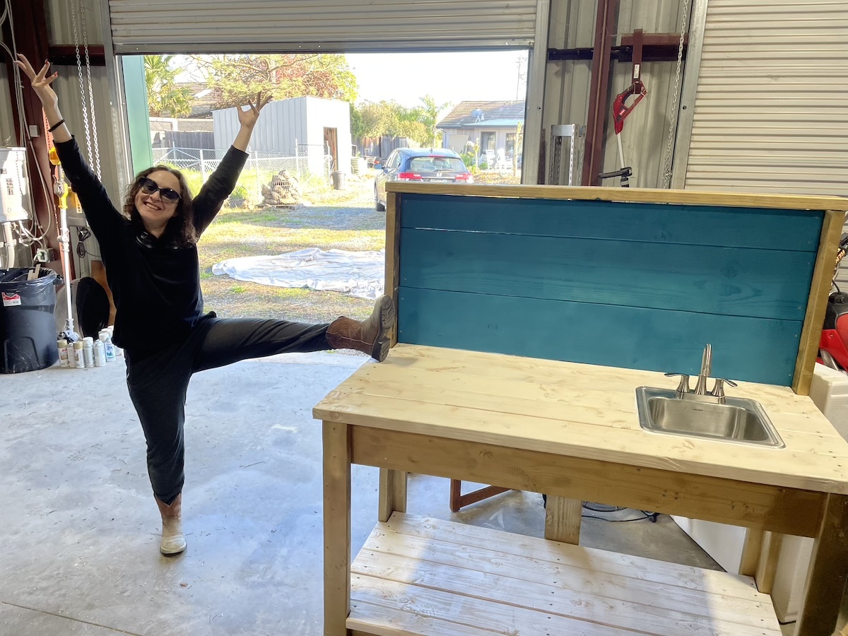 Delphine in casual clothes - long black shirt and black sweatpants. She is wearing cowboy boots. She has her leg up on a white and blue and gold sink that she made. She is in a woodworking shop with a cement floor. Her hands are up in the air in celebration.