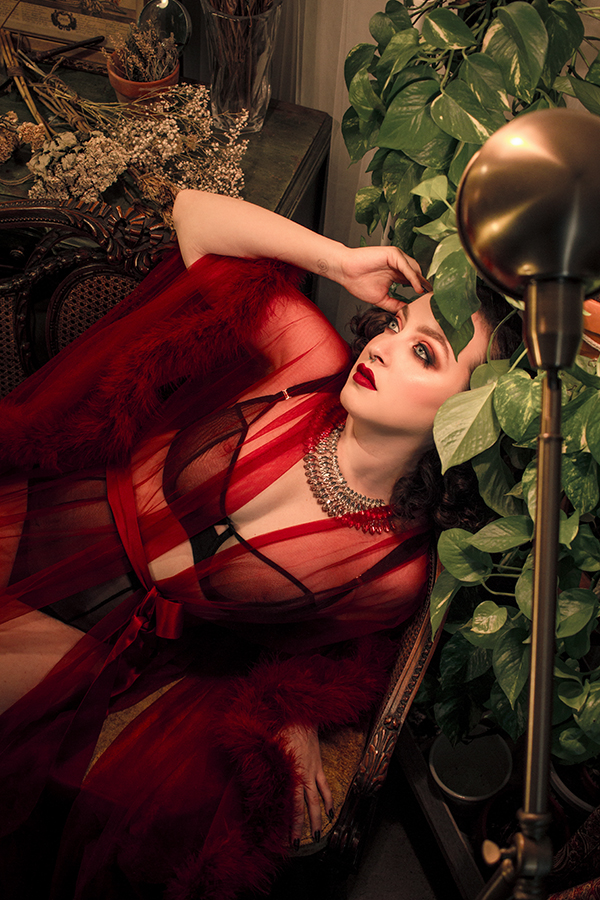 Delphine in a see through red robe surrounded by plants. She is wearing a large crystal necklace. The scene is quite dramatic and old-hollywood style. She is reclining.