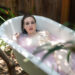 Delphine in a bathtub, she's wearing a white slip and the water is pink from a bath bomb. The tub is outdoors.